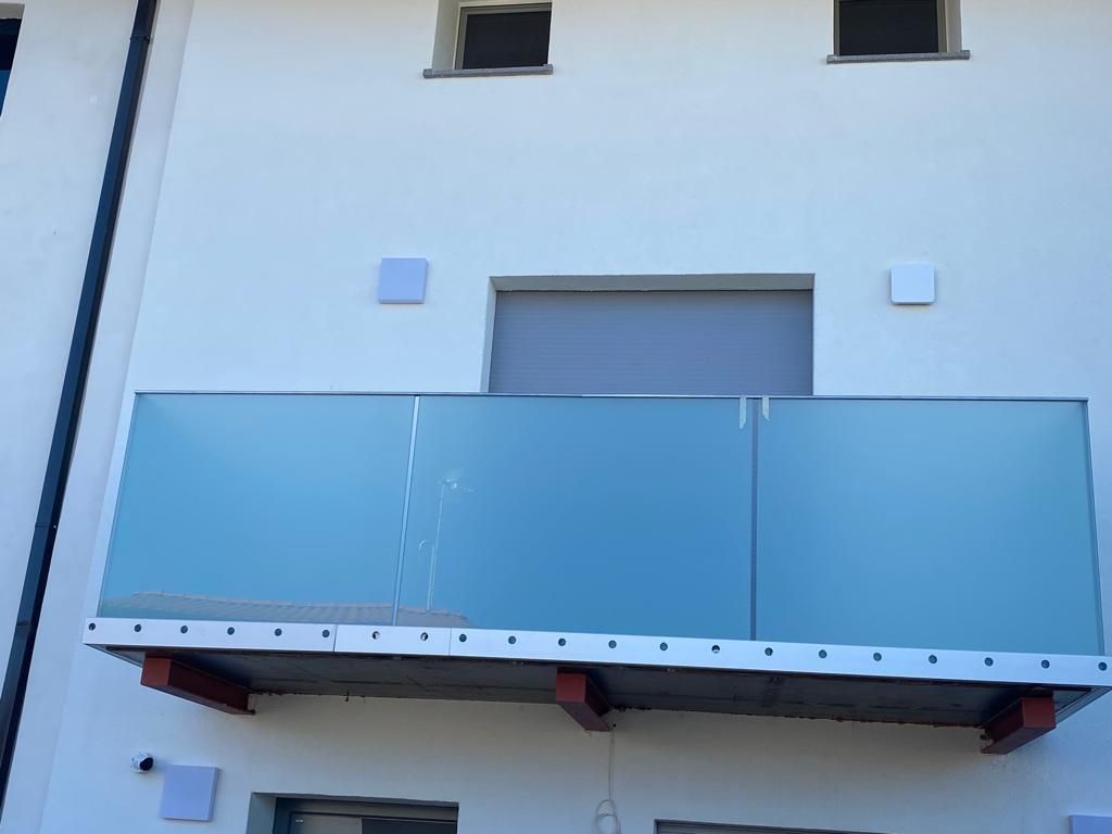 Esterno di un edificio bianco con balcone dotato di pannelli in vetro satinato e un bordo di rifinitura metallica parzialmente installato.