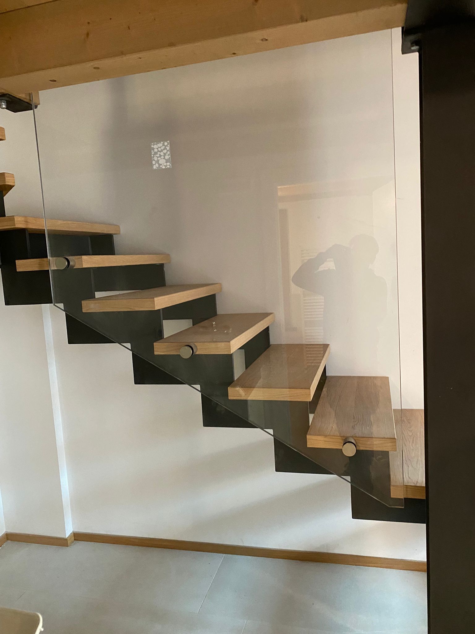 In una stanza minimalista, una scala sospesa con gradini in legno chiaro e supporti in metallo scuro si erge dietro una parete divisoria in vetro.
