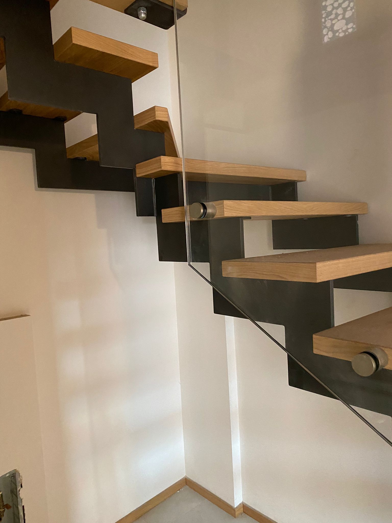 Scala moderna sospesa con gradini in legno chiaro e struttura in metallo grigio scuro su una parete bianca.