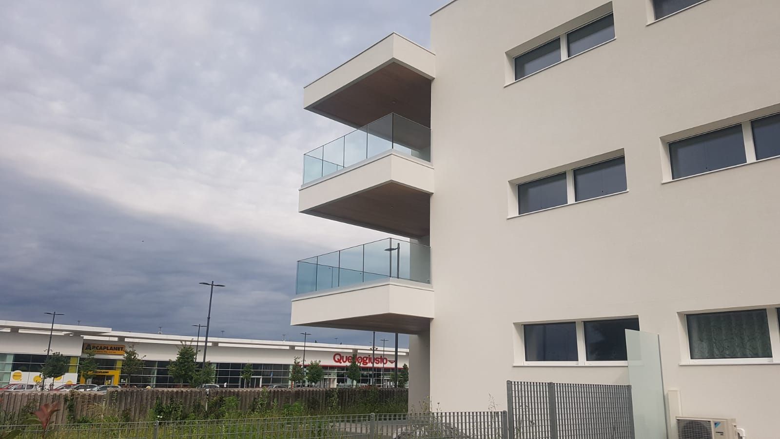 Moderno condominio dai colori chiari con balconi con ringhiere in vetro, stagliato contro un cielo nuvoloso, vicino a una zona commerciale.