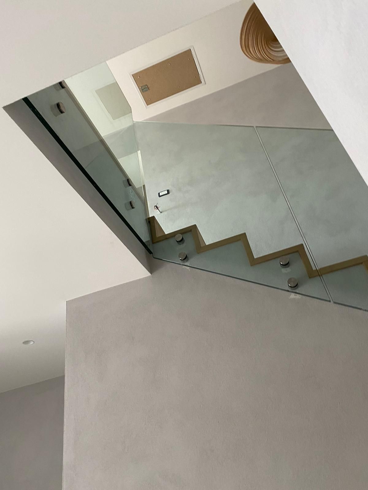 Vista dal basso di una scala interna con ringhiere in vetro, pareti color cemento e un pannello a incasso nel soffitto.