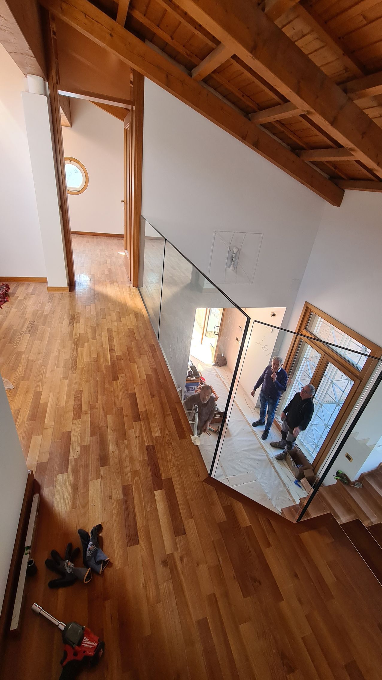 Inquadratura dall'alto di un corridoio con pavimento in legno e ringhiera in vetro che si affaccia su un piano inferiore, con due figure in primo piano.
