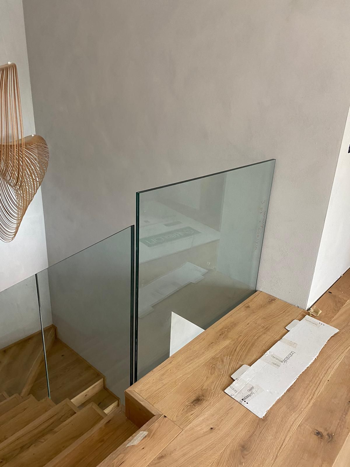 Una scala con gradini in legno chiaro e ringhiere in vetro, vista da un pianerottolo con una parete color bianco sporco dalla texture particolare.