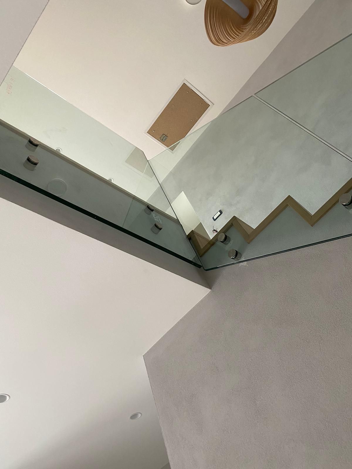 Una vista dal basso di un interno moderno caratterizzato da ringhiere delle scale in vetro e una lampada a sospensione intrecciata su un soffitto bianco.