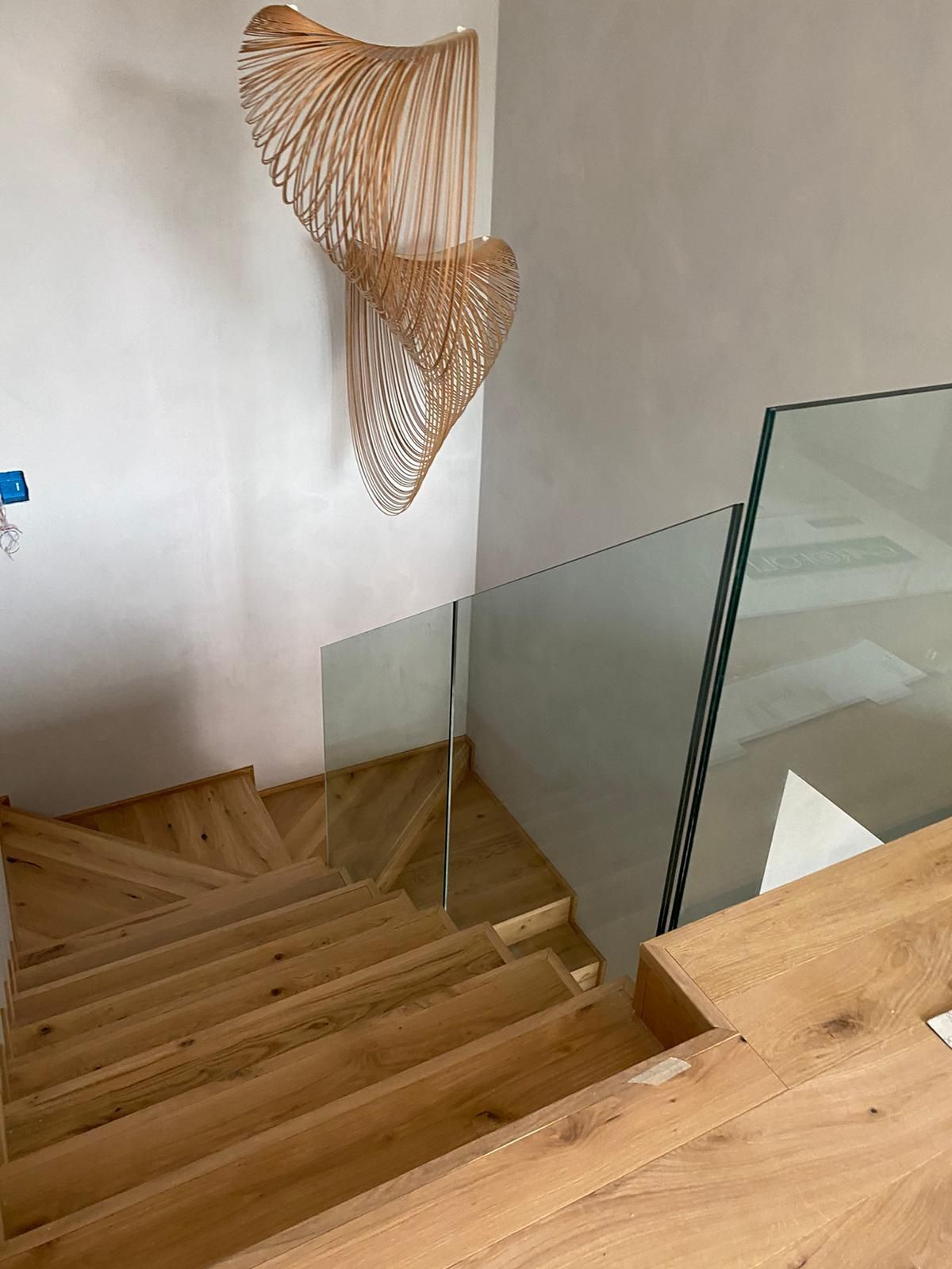 Una vista dall'alto di una scala in legno con ringhiera in vetro e una lampada scultorea intrecciata appesa alla parete.