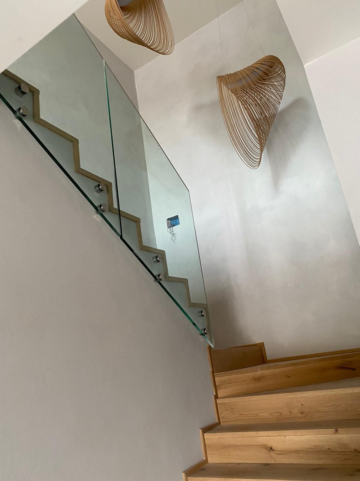 Una scala in legno con ringhiera in vetro e supporti distanziatori, illuminata da due lampade a sospensione intrecciate.
