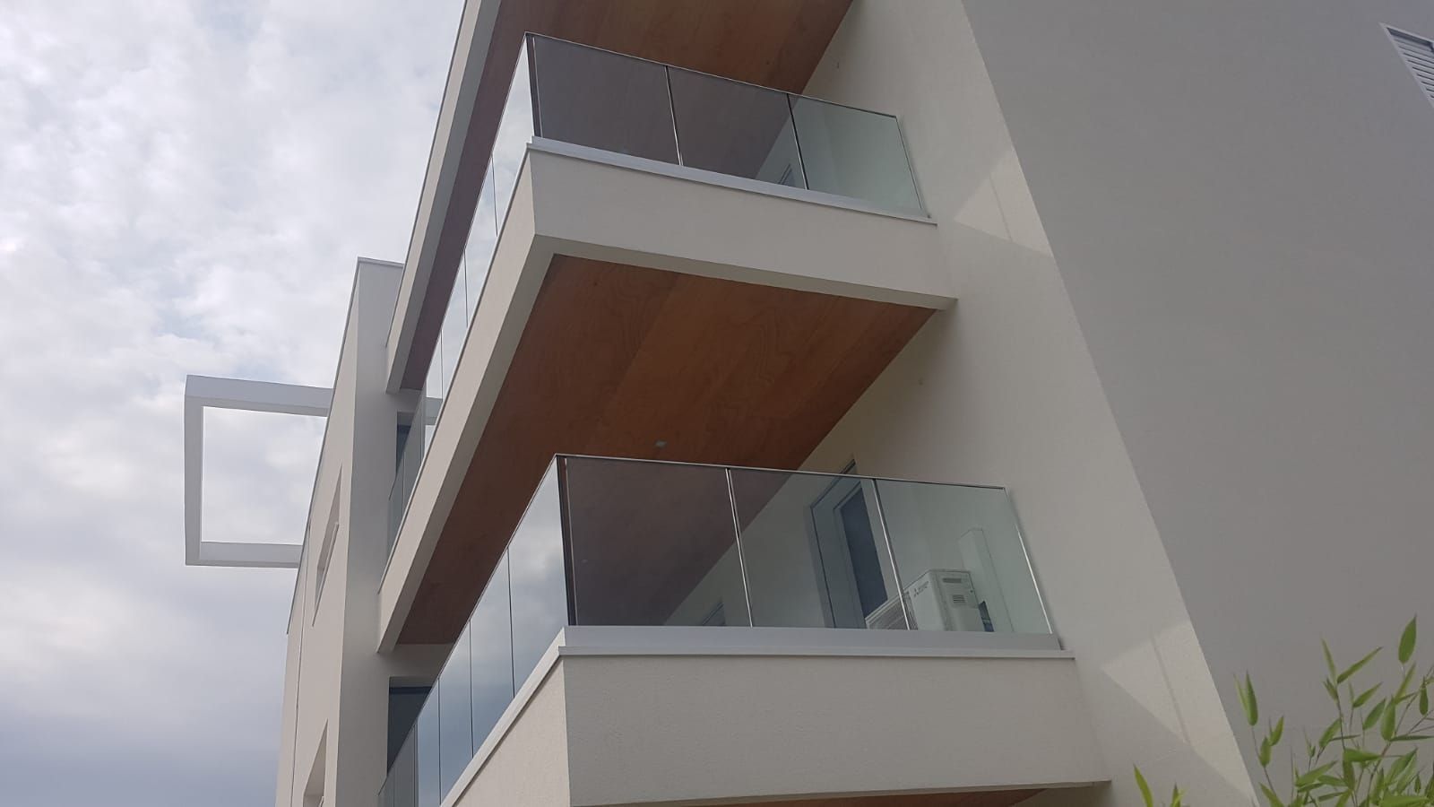 Vista dal basso della facciata di un edificio moderno, caratterizzata da balconi sovrapposti con ringhiere in vetro e soffitti rivestiti in legno.