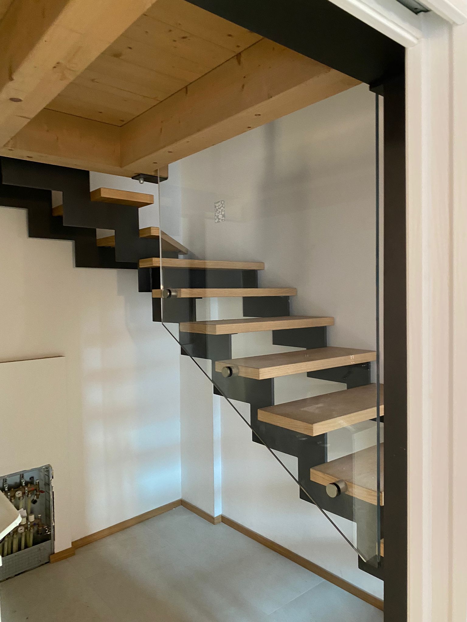 Scala moderna sospesa con gradini in legno e struttura in metallo nero, caratterizzata da una ringhiera con pannello in vetro su un lato.