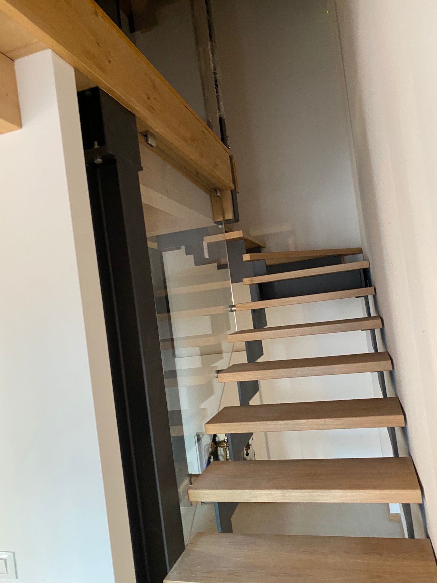 Scala moderna in legno con gradini a vista, struttura in metallo scuro e ringhiera in vetro, su uno sfondo di parete bianca.