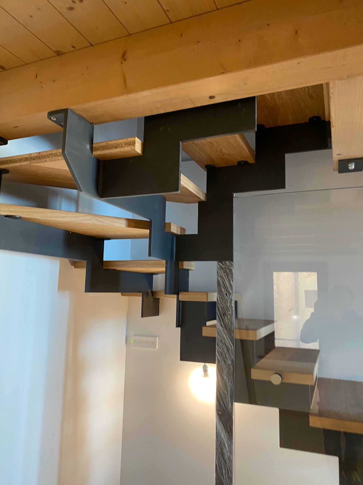 Scala con montanti a zig-zag in acciaio scuro che sorreggono gradini in legno e un montante verticale decorativo con motivi ornamentali.