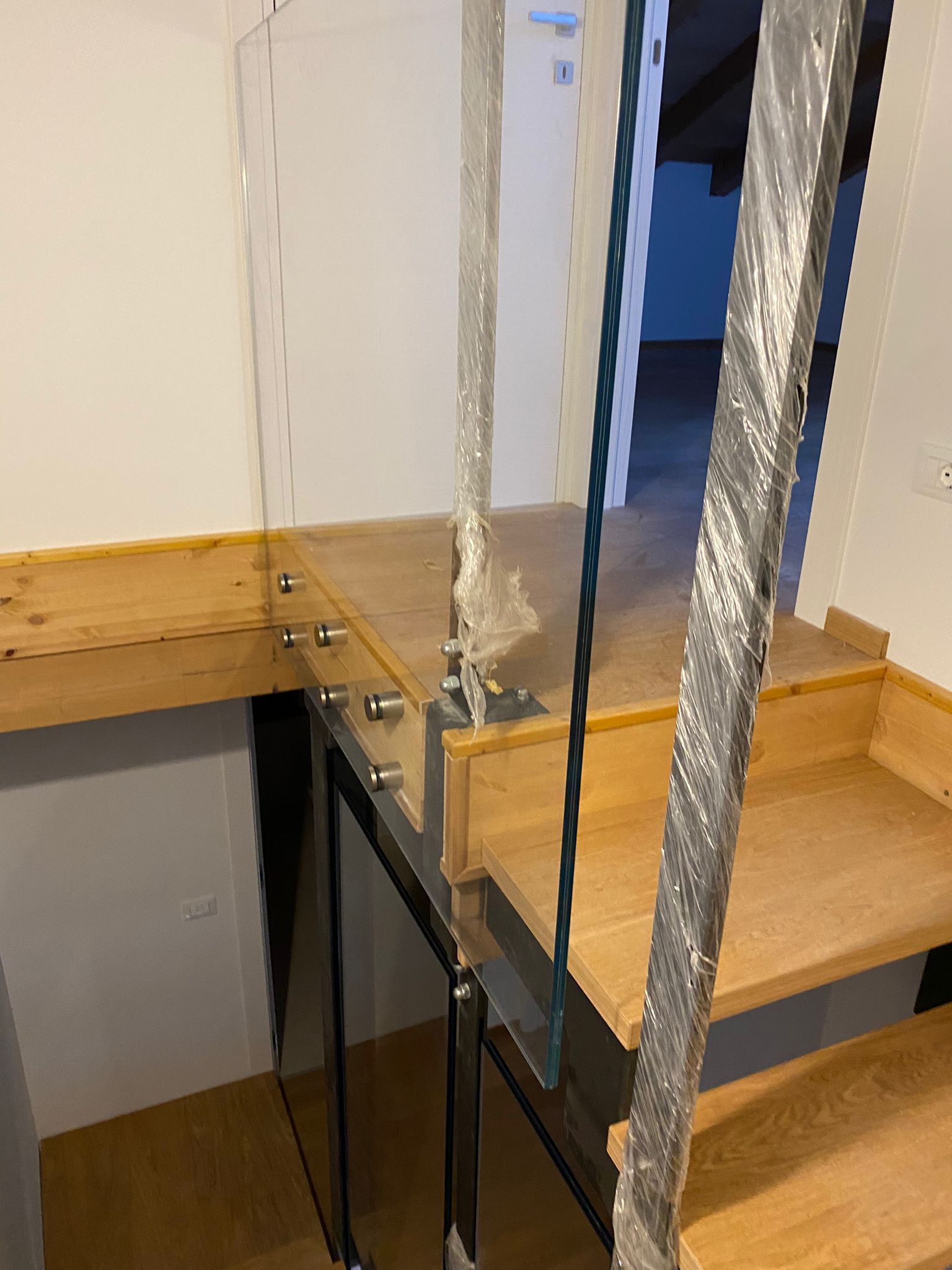 Una scala in legno con ringhiera in vetro e fissaggi laterali in metallo, parzialmente ricoperta da un involucro protettivo in plastica.