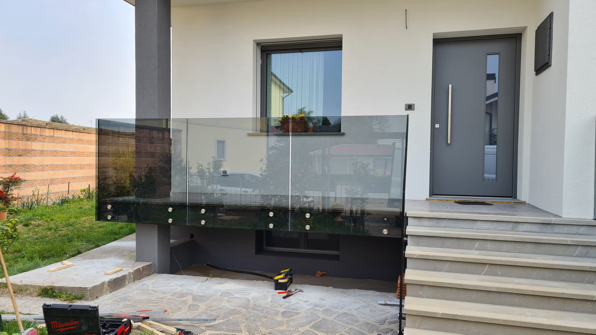 Una ringhiera in vetro per balcone annessa a una casa moderna con scalini in pietra grigia e porta d'ingresso.