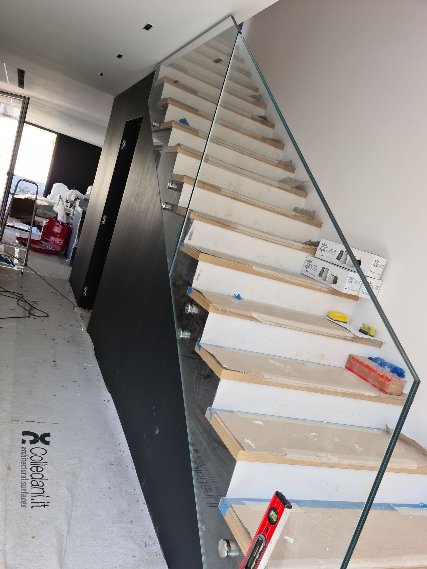 Scala in costruzione con gradini in legno, ringhiera in vetro e parete d'accento scura in una casa moderna.