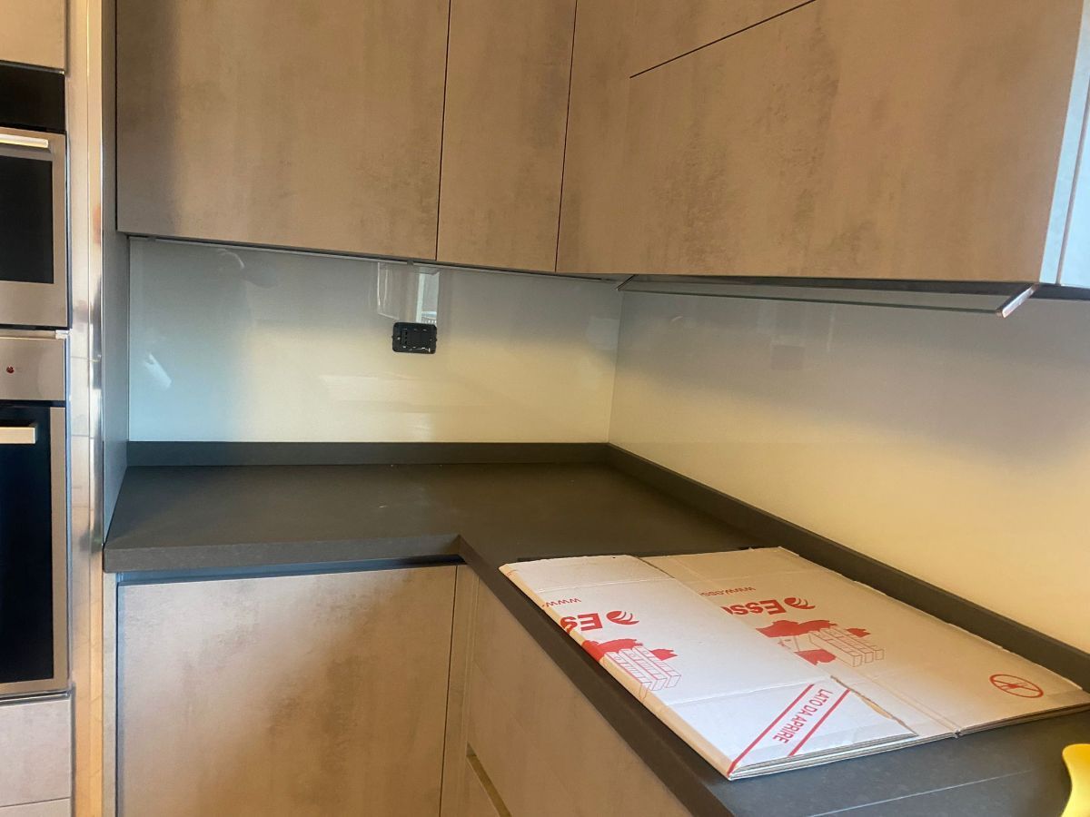 Un angolo cucina caratterizzato da mobili con finitura in legno chiaro, un piano di lavoro grigio scuro e un paraschizzi in vetro bianco pulito.