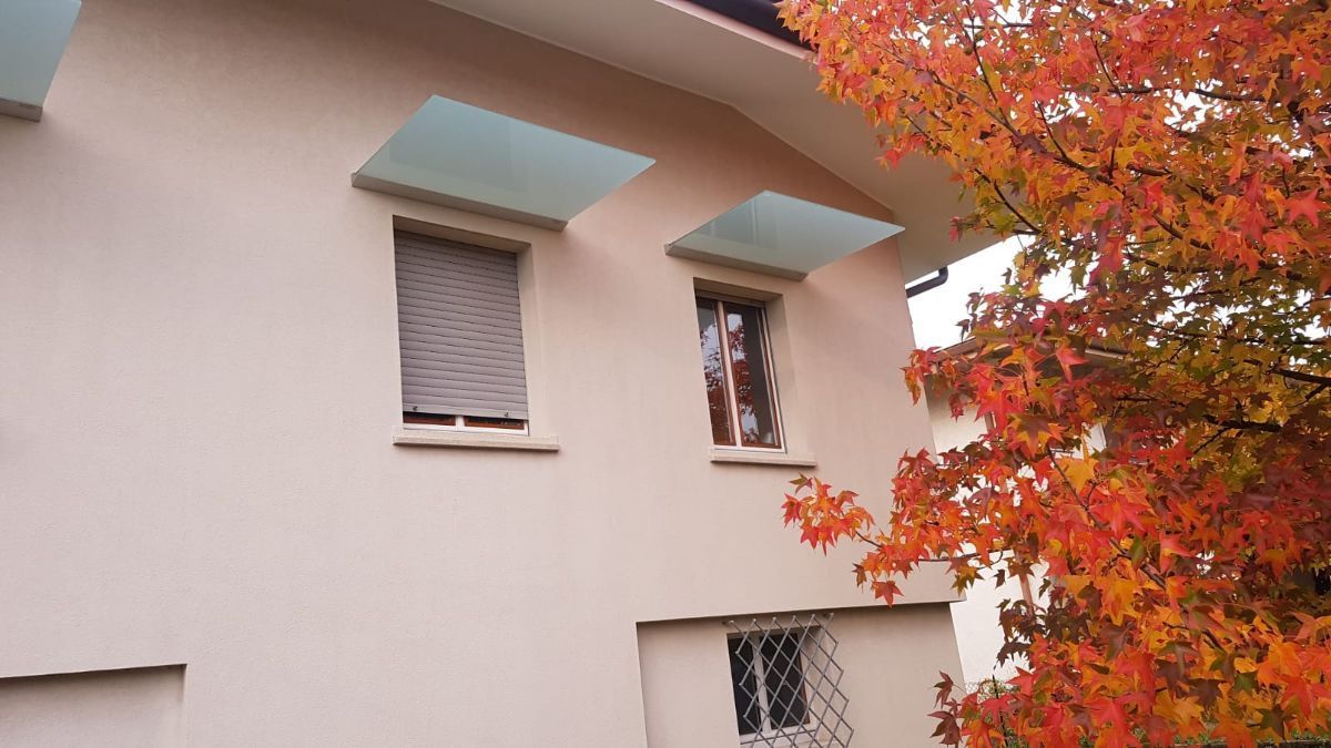 Esterno di una casa color pesca con due finestre al piano superiore dotate di tende da sole in vetro e una finestra al piano inferiore, accanto a un albero autunnale.