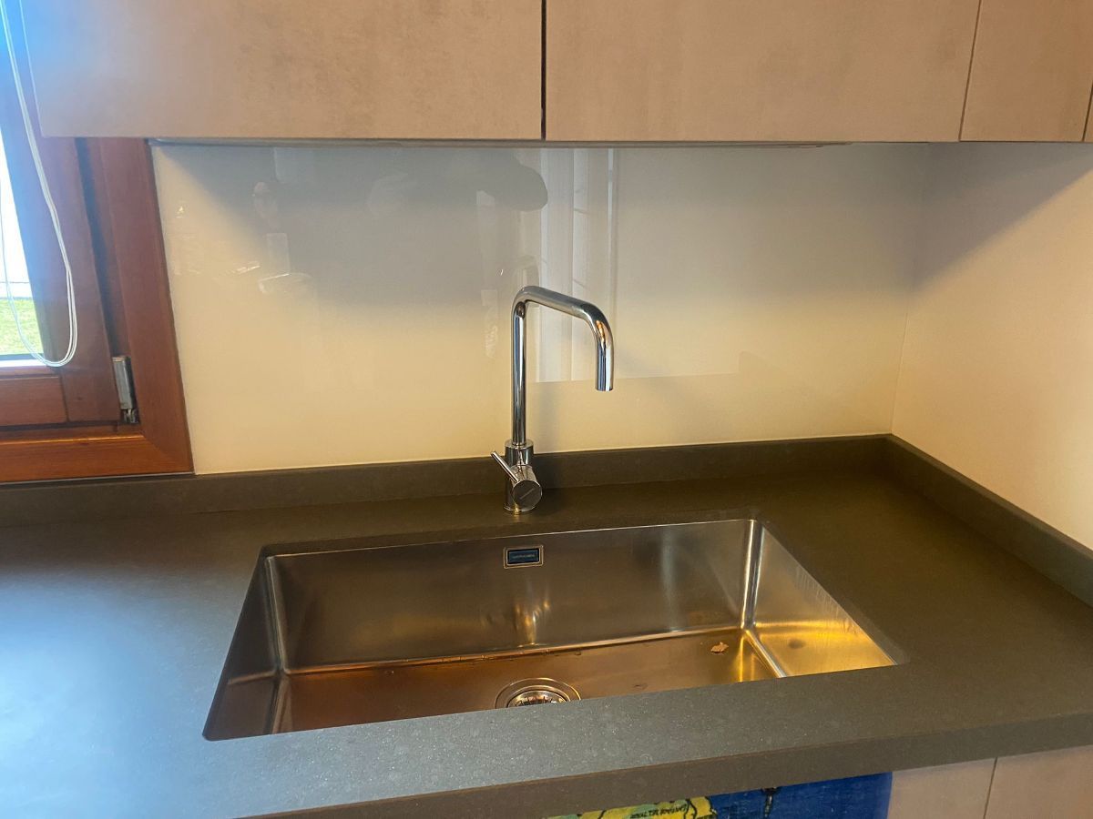 Un lavello da cucina in acciaio inox con rubinetto cromato, incastonato in un piano di lavoro grigio scuro su un paraschizzi bianco liscio.