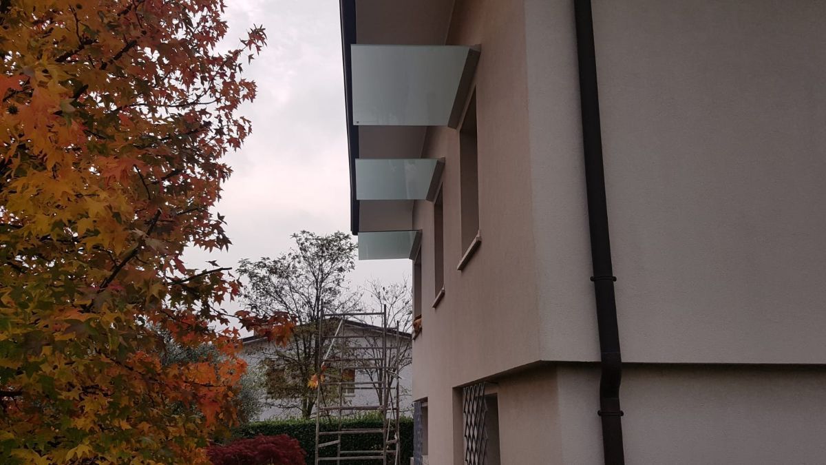 Vista laterale di una casa beige con diverse sezioni di tetto sporgenti, un tubo di scarico scuro e alberi autunnali sullo sfondo.