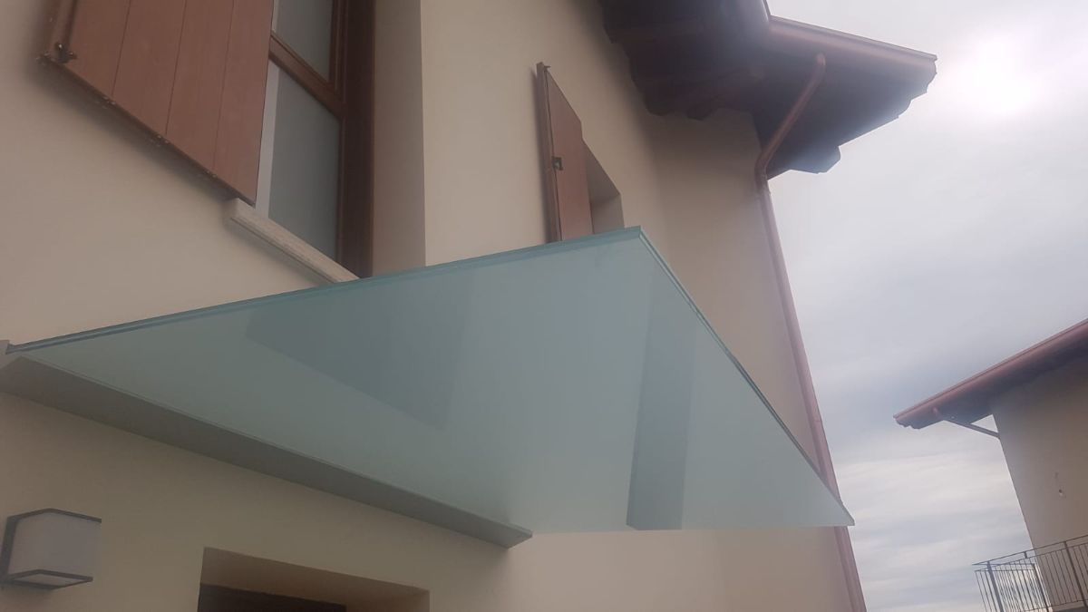 Una moderna tettoia in vetro satinato, installata sopra la porta d'ingresso di una casa color sabbia con persiane marroni, sotto un cielo nuvoloso.