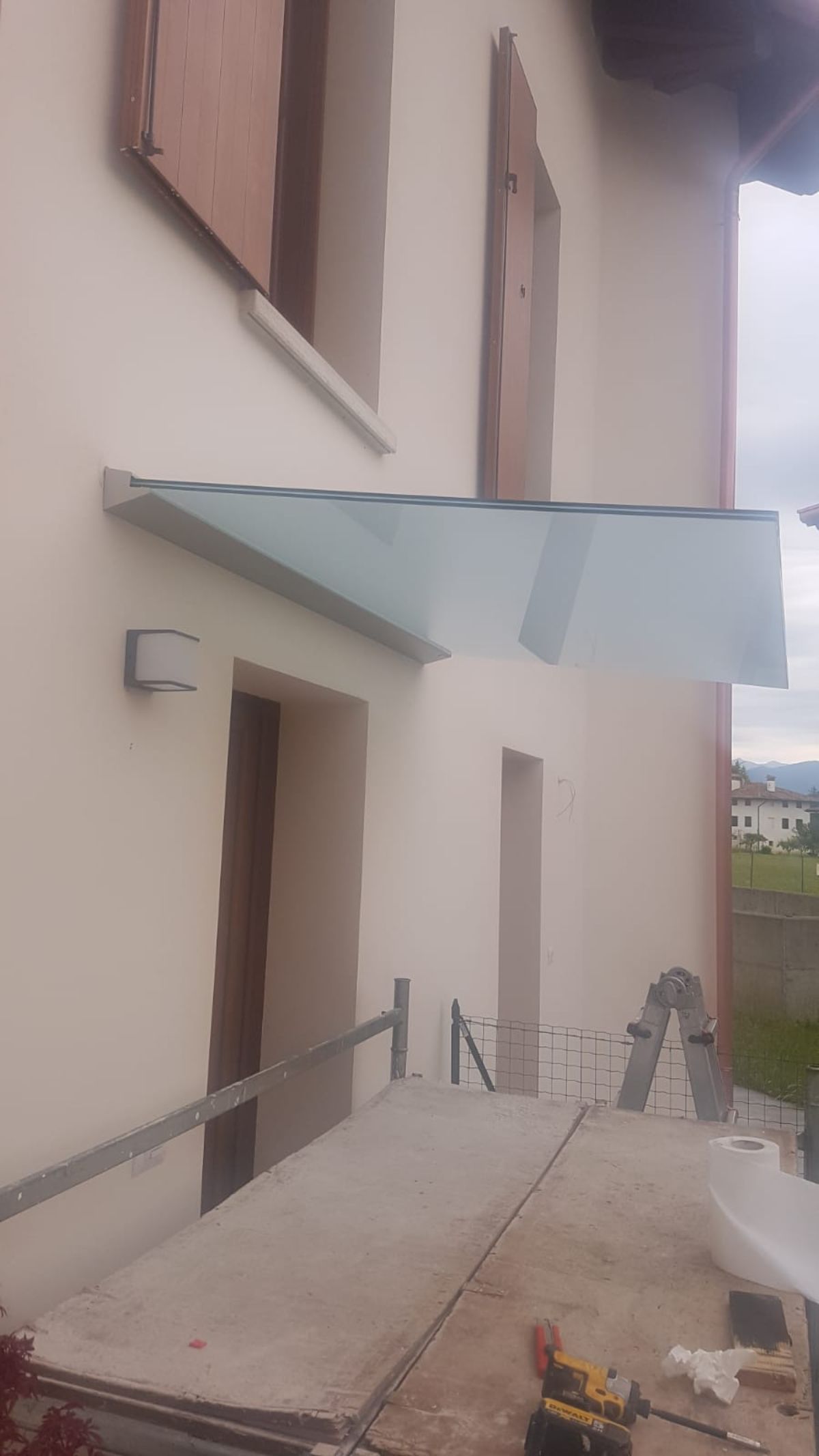 Una pensilina in vetro installata sopra una porta sulla facciata bianca di un edificio, con impalcature e attrezzi da costruzione nelle vicinanze.