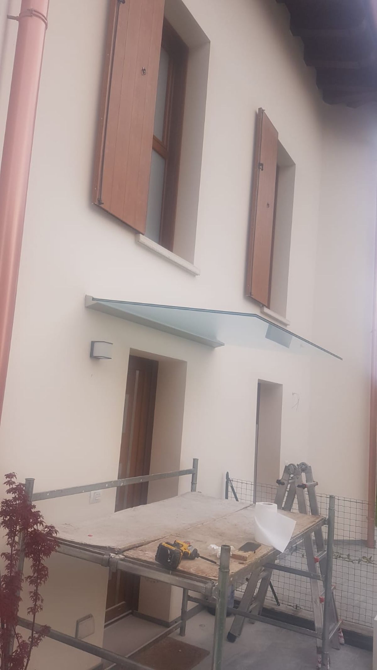 Esterno di un edificio dai colori tenui con persiane in legno alle finestre superiori e una moderna tettoia in vetro sopra un ingresso e un'impalcatura.