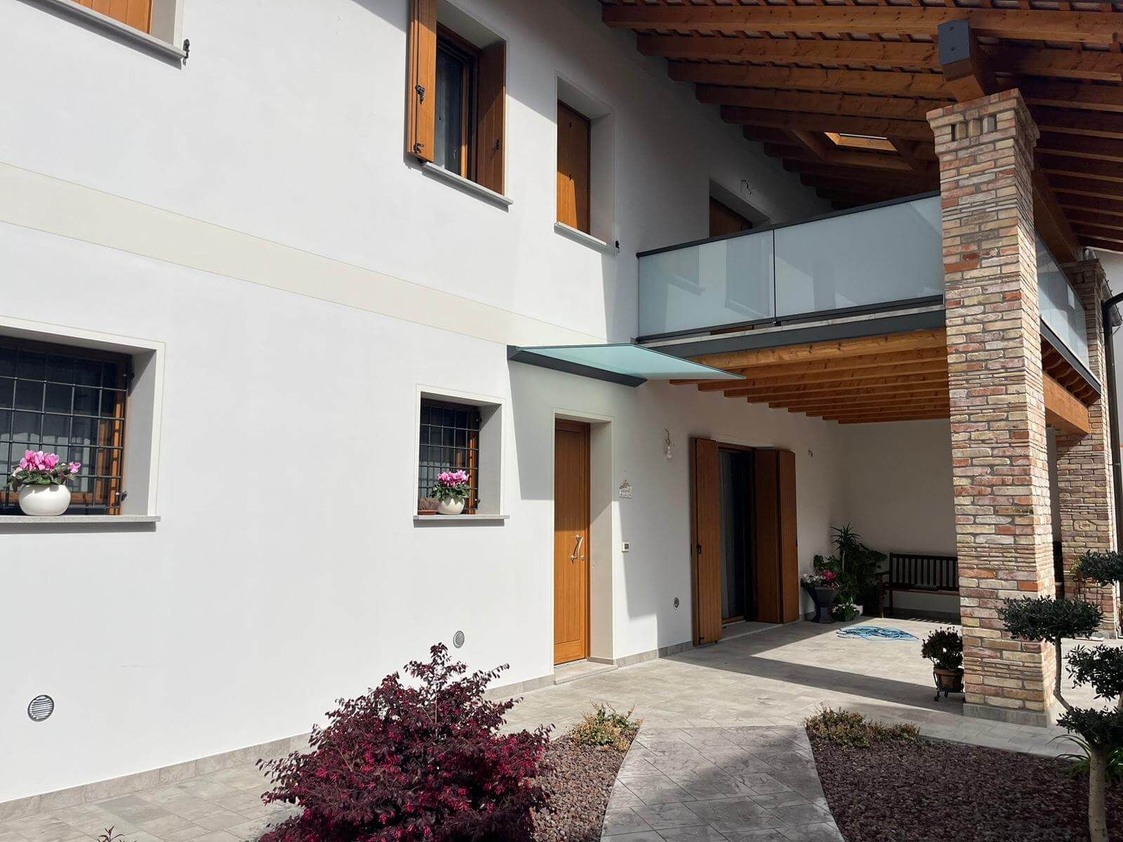 Una casa bianca a due piani con persiane in legno, un pilastro in pietra, un balcone e un ingresso con patio.
