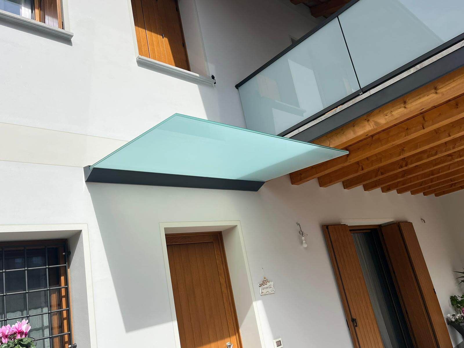 Sopra una porta d'ingresso in legno di un edificio bianco è installata una tettoia in vetro, con un balcone visibile al di sopra.