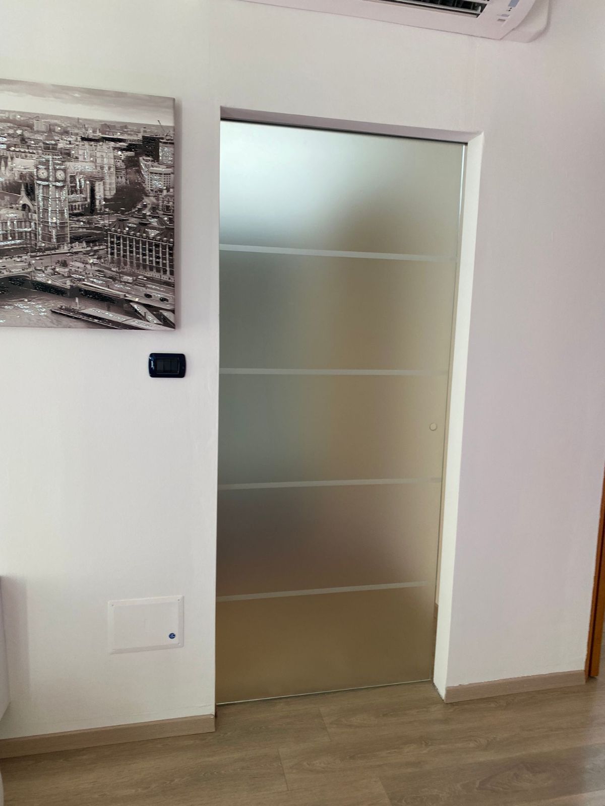 Una porta in vetro satinato con cinque pannelli orizzontali installata in una parete bianca accanto a un'opera d'arte raffigurante lo skyline di una città.