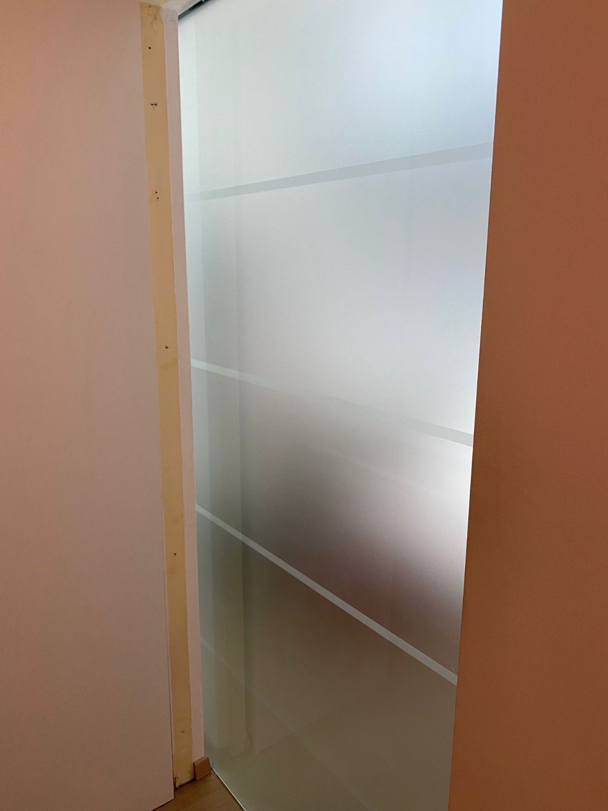Una porta in vetro satinato con strisce orizzontali, incorniciata da una modanatura in legno chiaro, su una parete liscia.