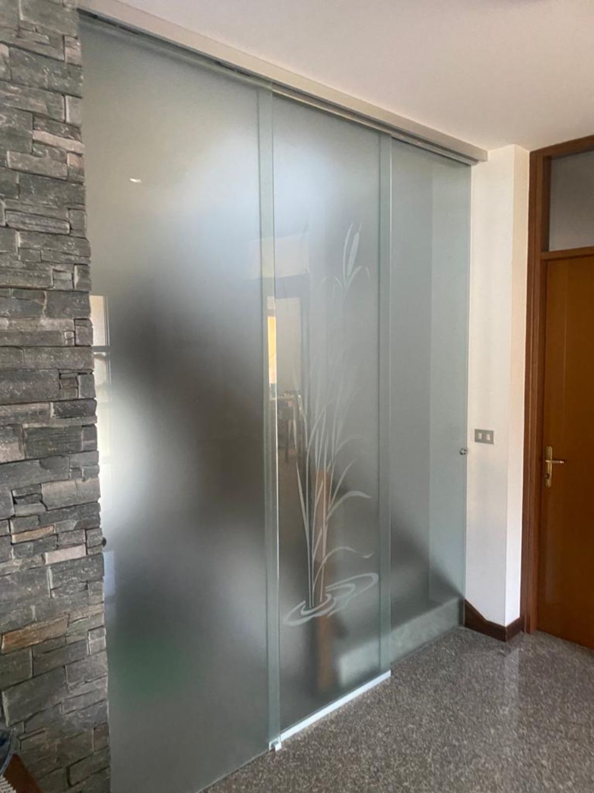 Una porta scorrevole in vetro con finitura satinata e motivo a canne incise, inserita tra un muro in pietra e una porta in legno.