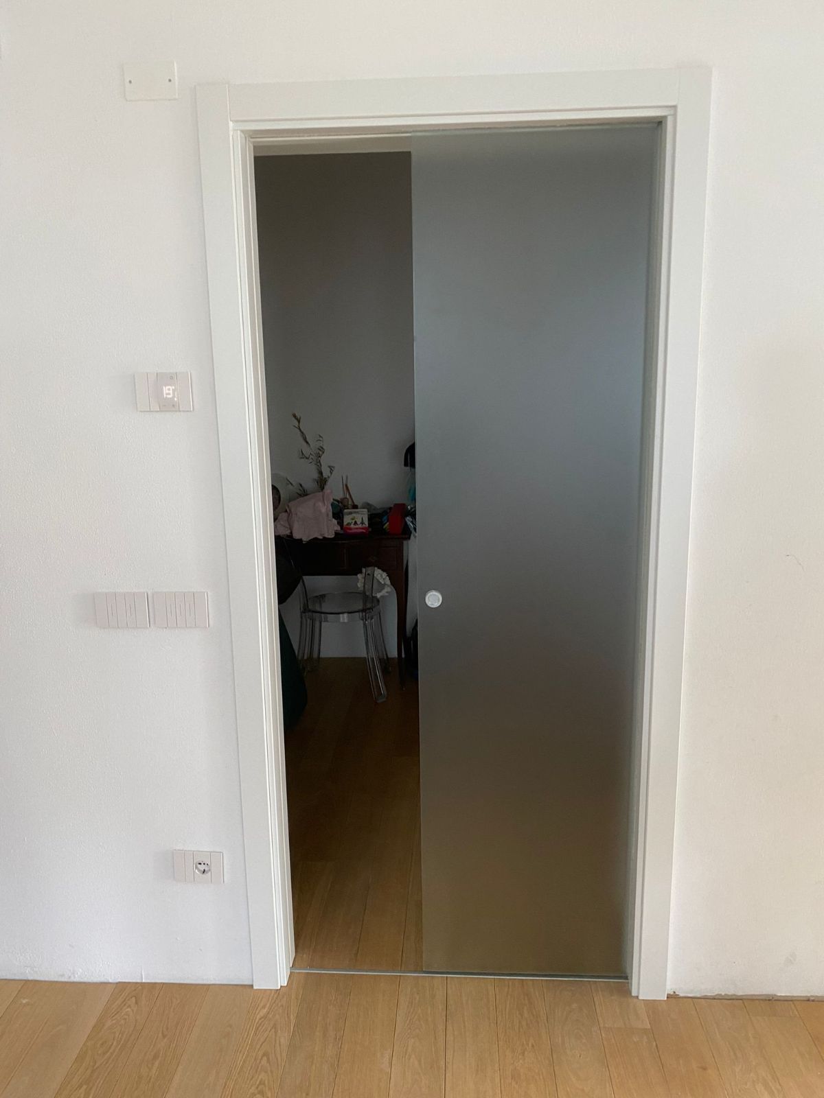 Una porta scorrevole in vetro satinato semitrasparente, incastonata in una parete bianca, conduce a una stanza con pavimento in legno.