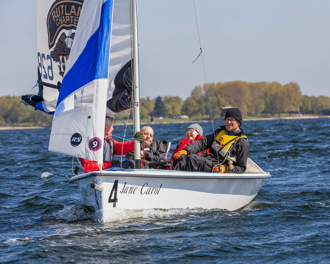 Small sailboat with three people racing on choppy blue water, white sail and flags raised