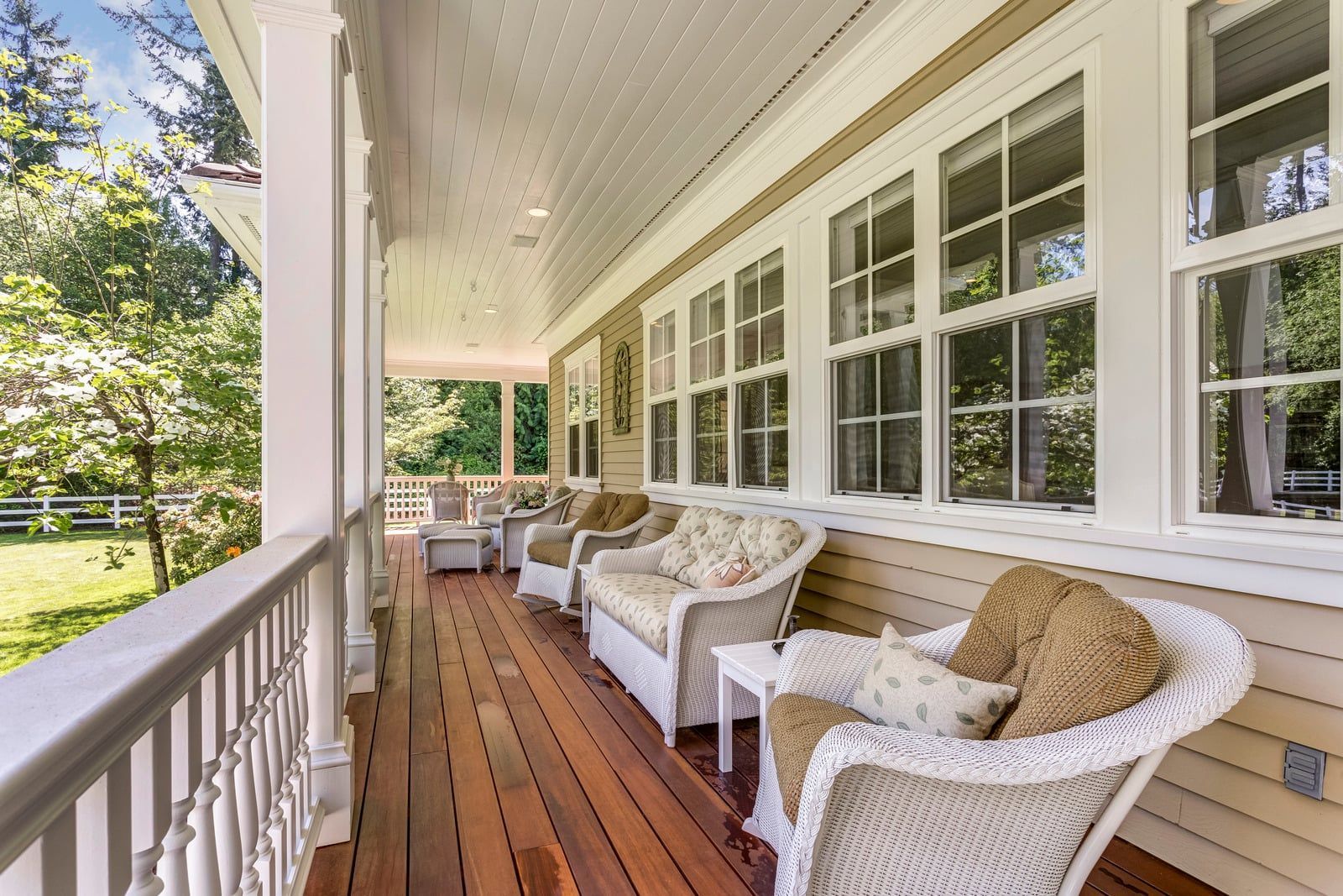 Covered front porch with white wicker chairs, wooden floor, and rows of windows along a beige house