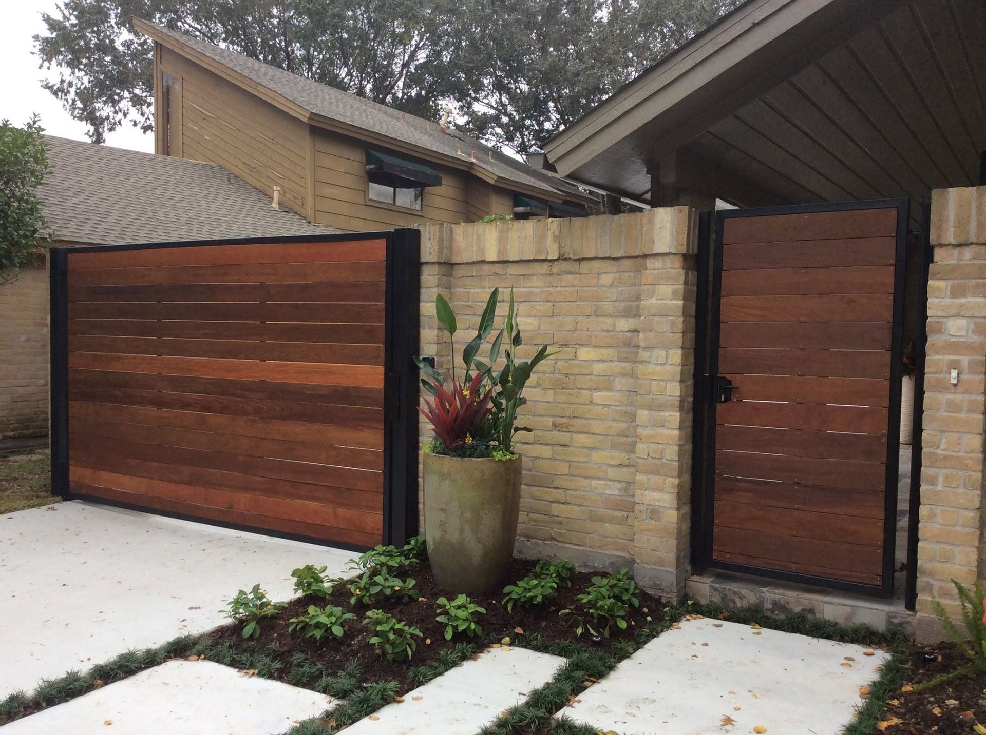 Modern house entrance with wood-and-stone gate, tall planter, and concrete walkway across the foreground