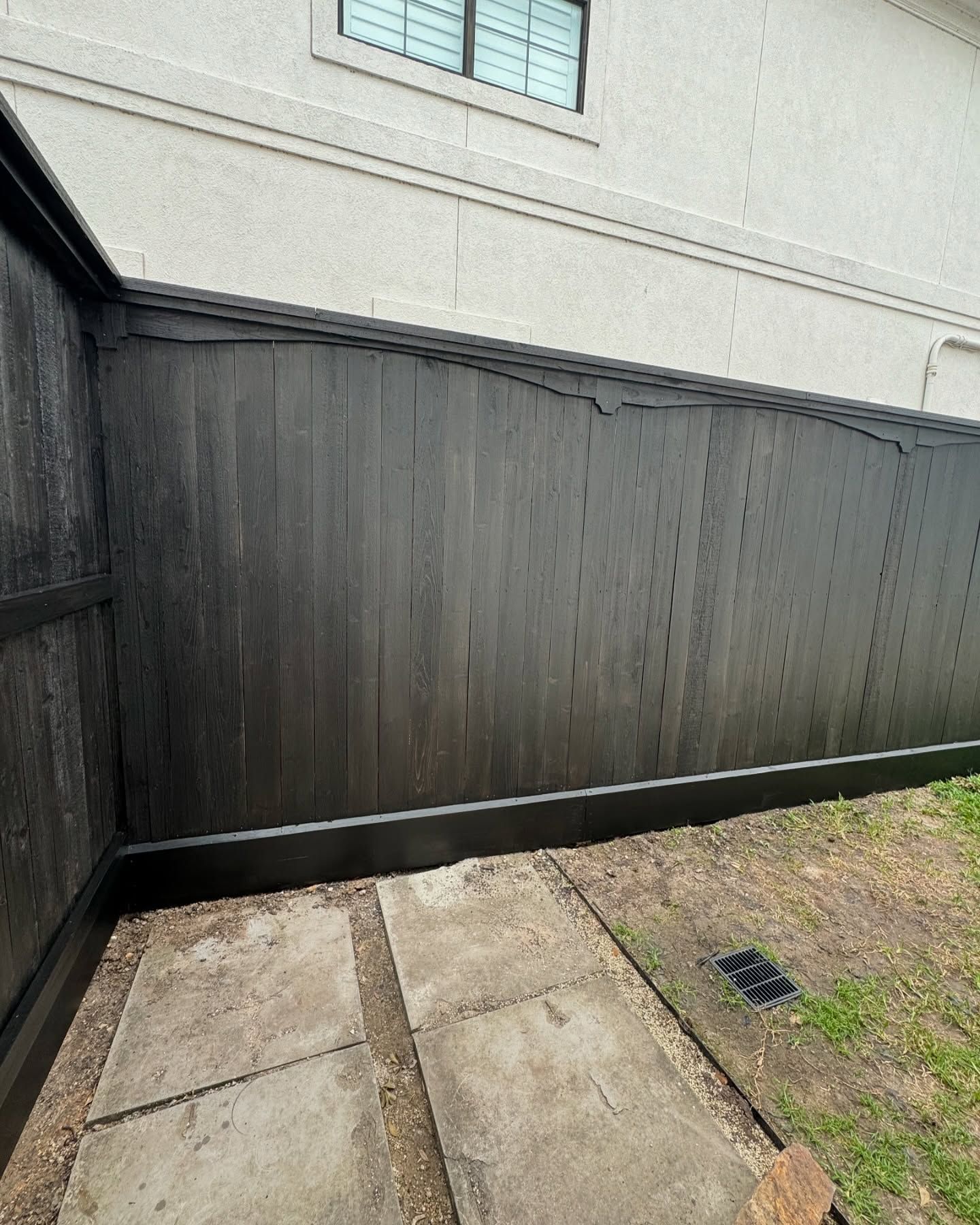 Black wooden fence beside a concrete walkway and patchy grass next to a light-colored building