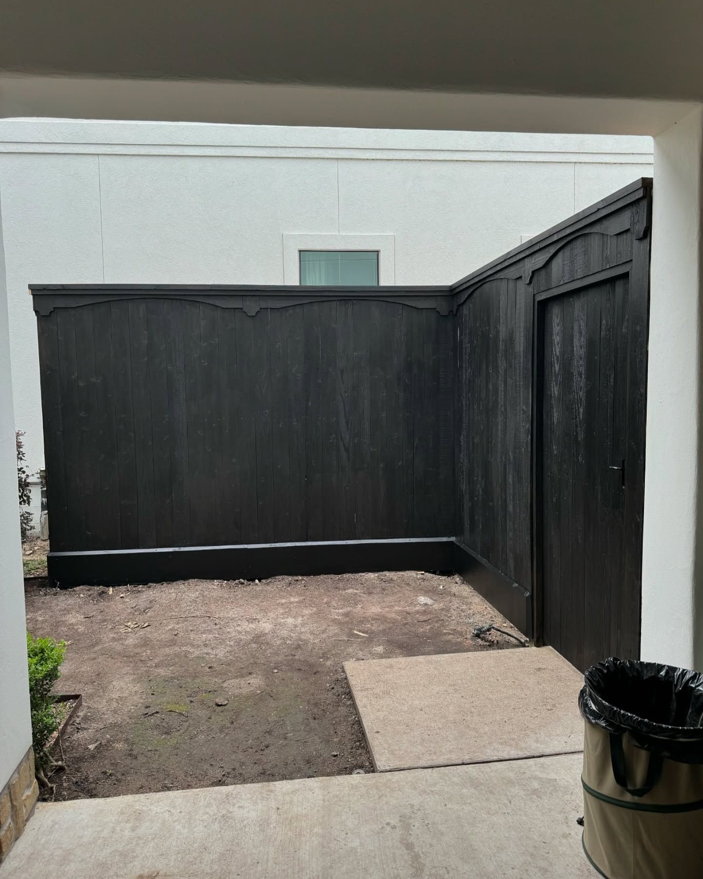 Small fenced courtyard with black wooden walls, bare ground, and a concrete step near a trash can.