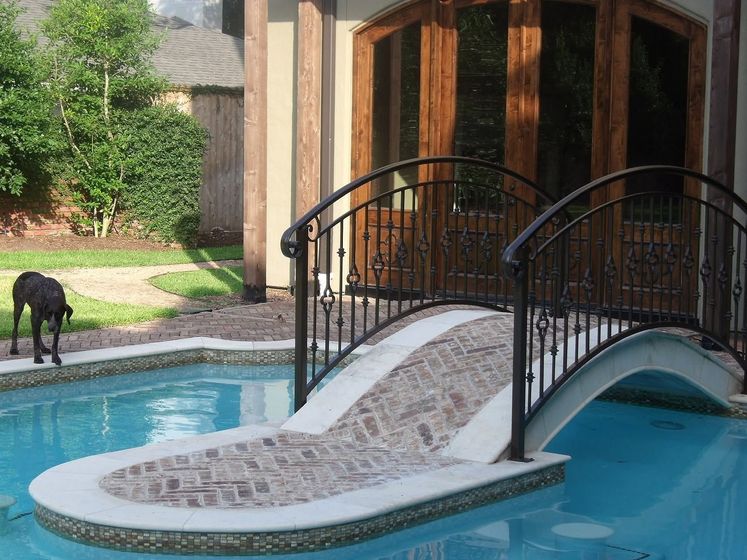 Stone bridge over a backyard pool beside a house, with a black dog nearby.