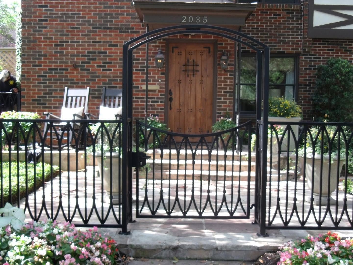 Brick house entrance with black wrought-iron gate, wooden front door, and flower beds