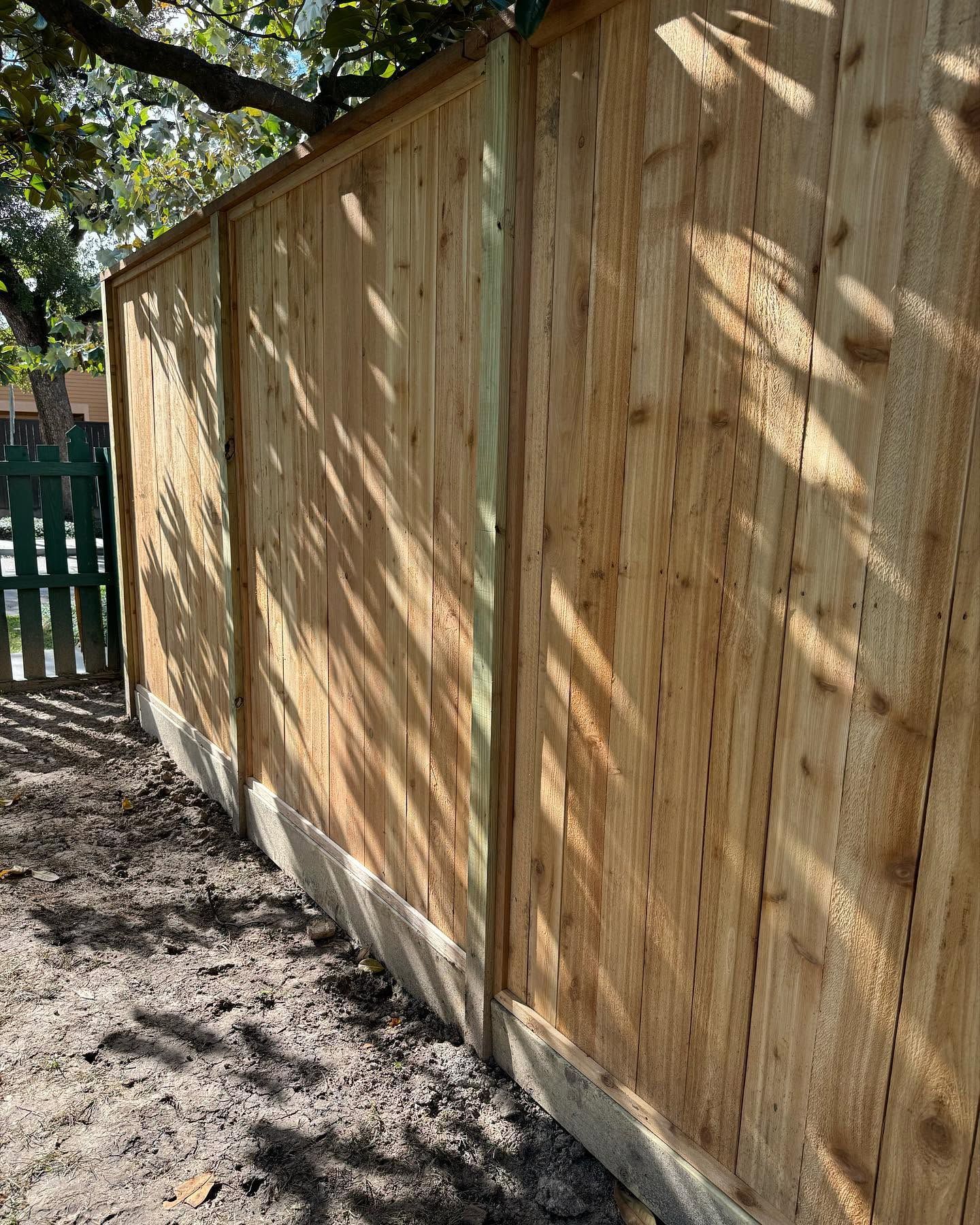 Wooden privacy fence along a dirt path, with sunlight and tree shadows.