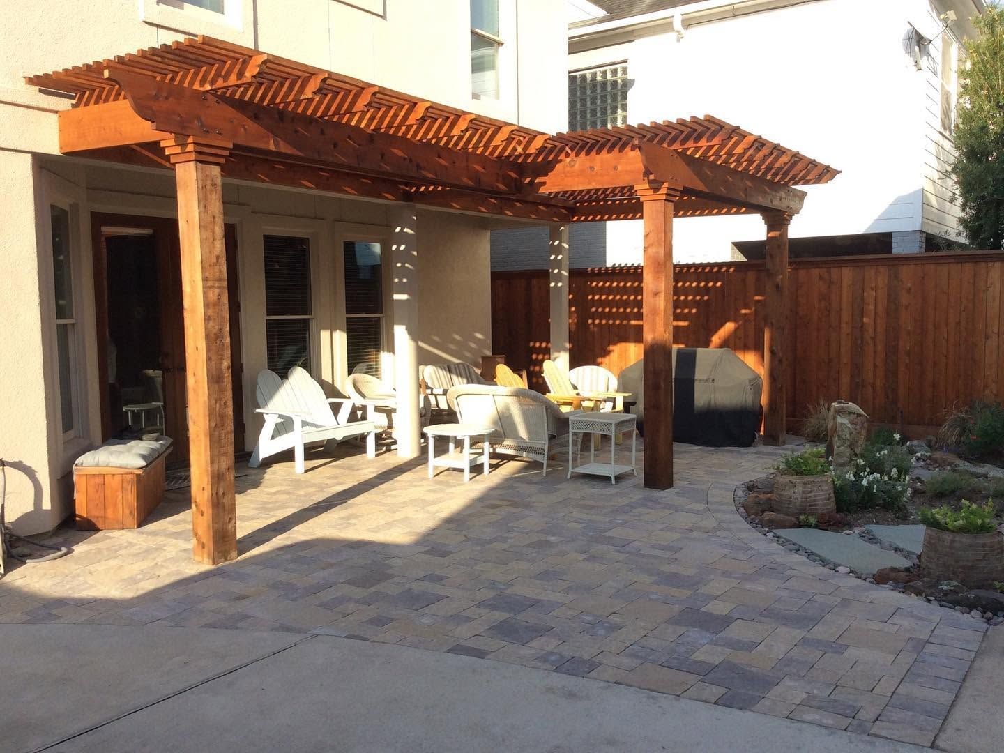 Patio with wooden pergola, white chairs, stone pavers, and a small garden beside a house