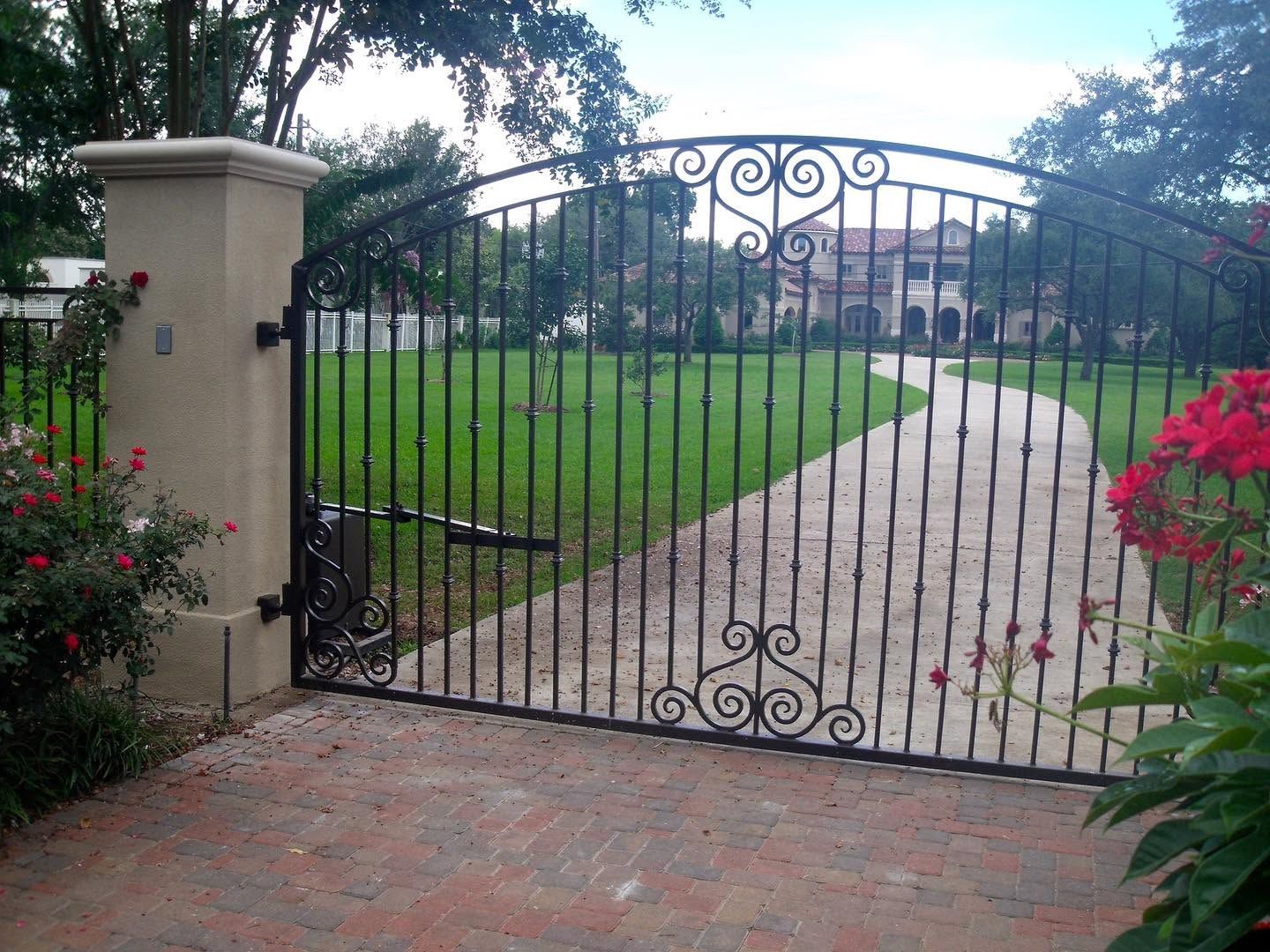 Wrought-iron gate opening to a curved driveway and green lawn beside a stone pillar and flowers.
