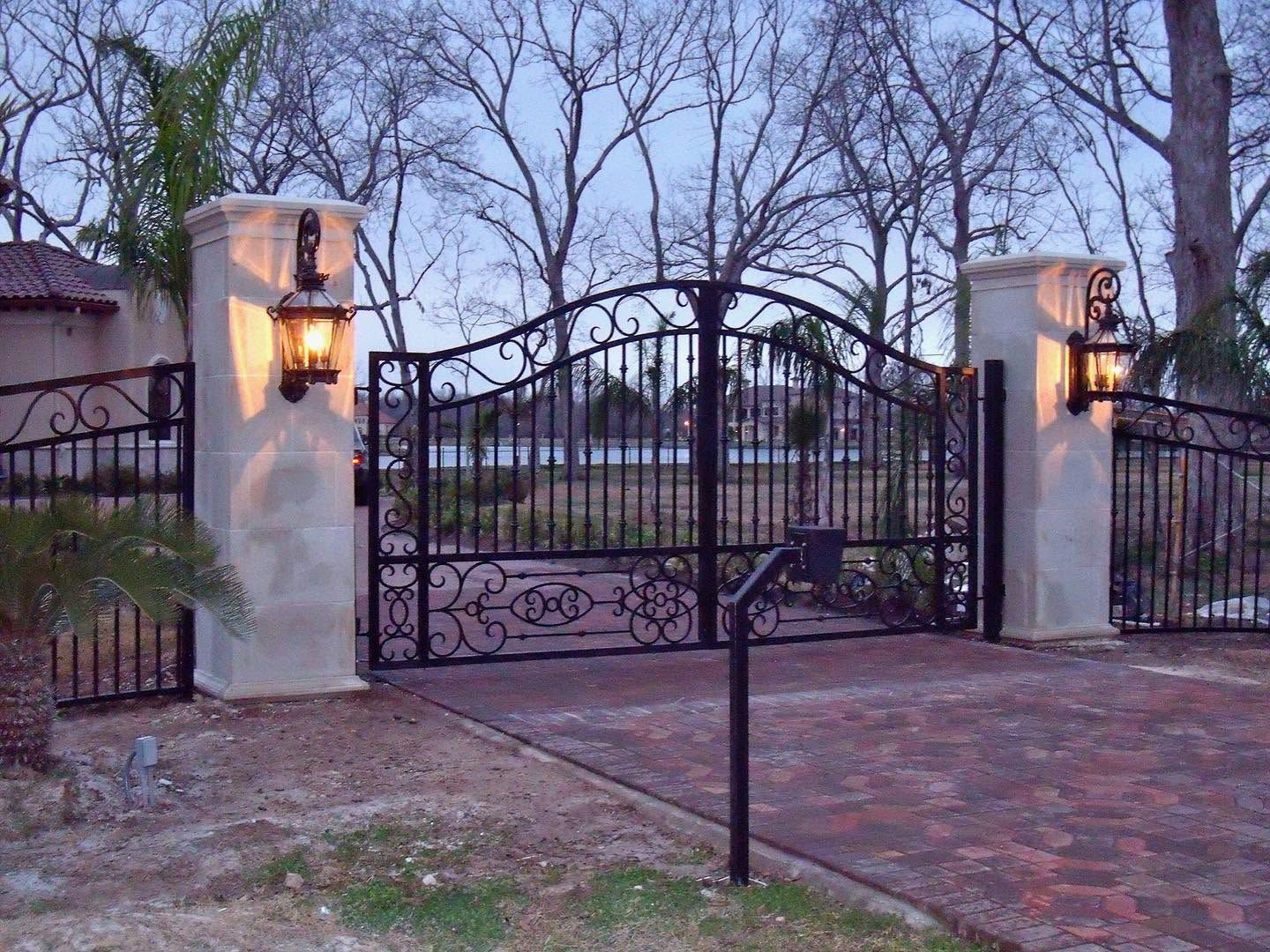 Black wrought-iron driveway gate between stone pillars with lanterns at dusk