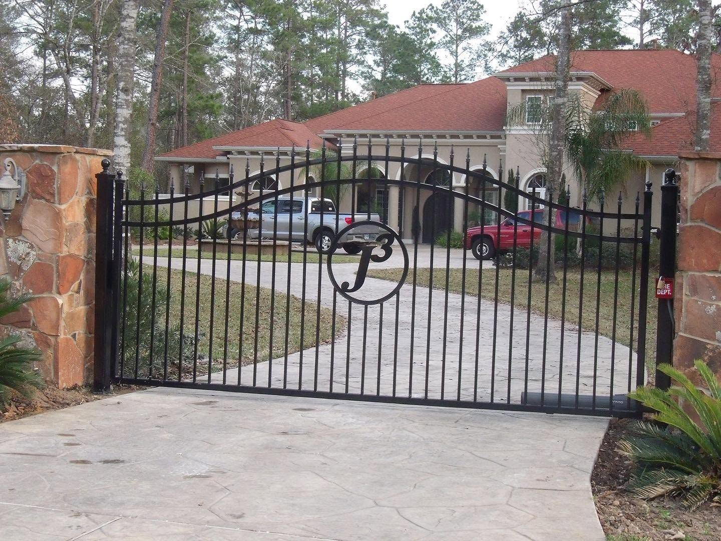 Black wrought-iron driveway gate in front of a red-roofed house and parked cars