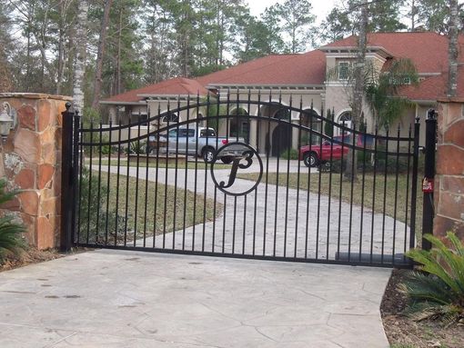 Black wrought-iron gate at driveway entrance to a red-roofed house with parked cars behind it