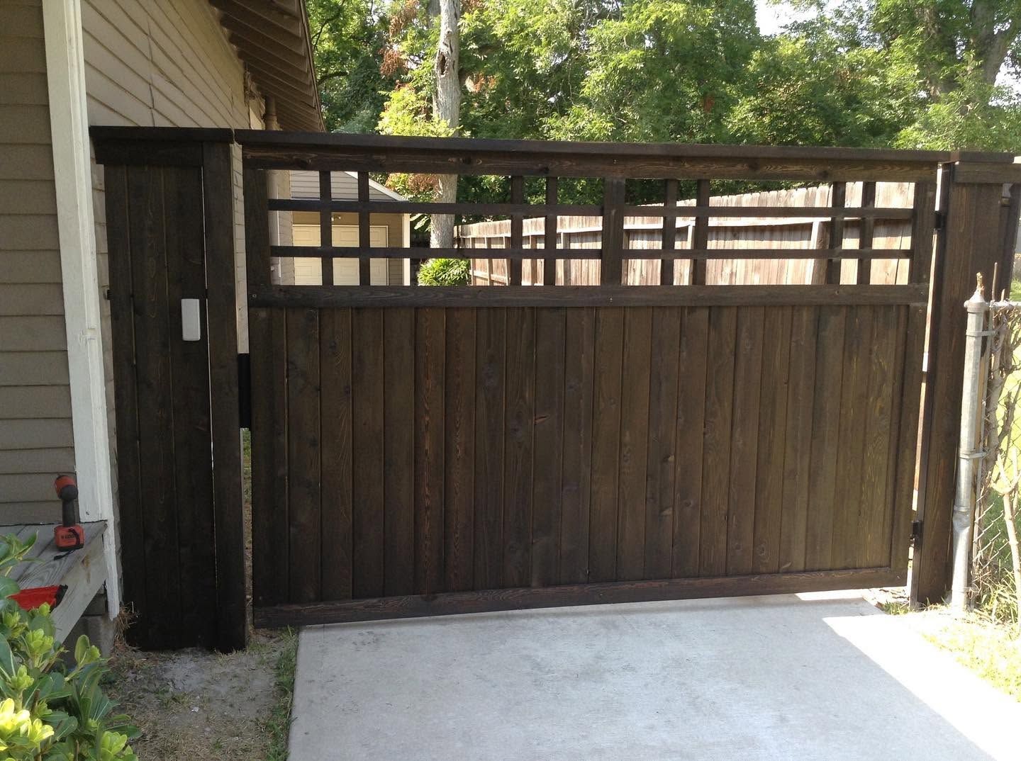 Dark wooden driveway gate with lattice top beside a house and concrete driveway