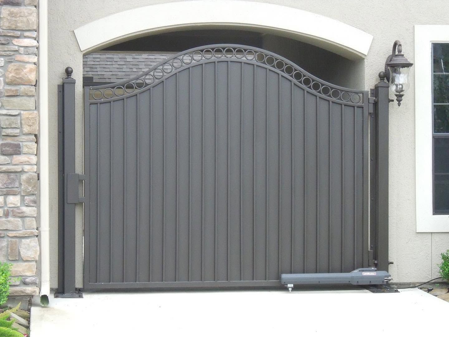 Gray metal driveway gate with decorative arch in front of a stone and stucco house exterior