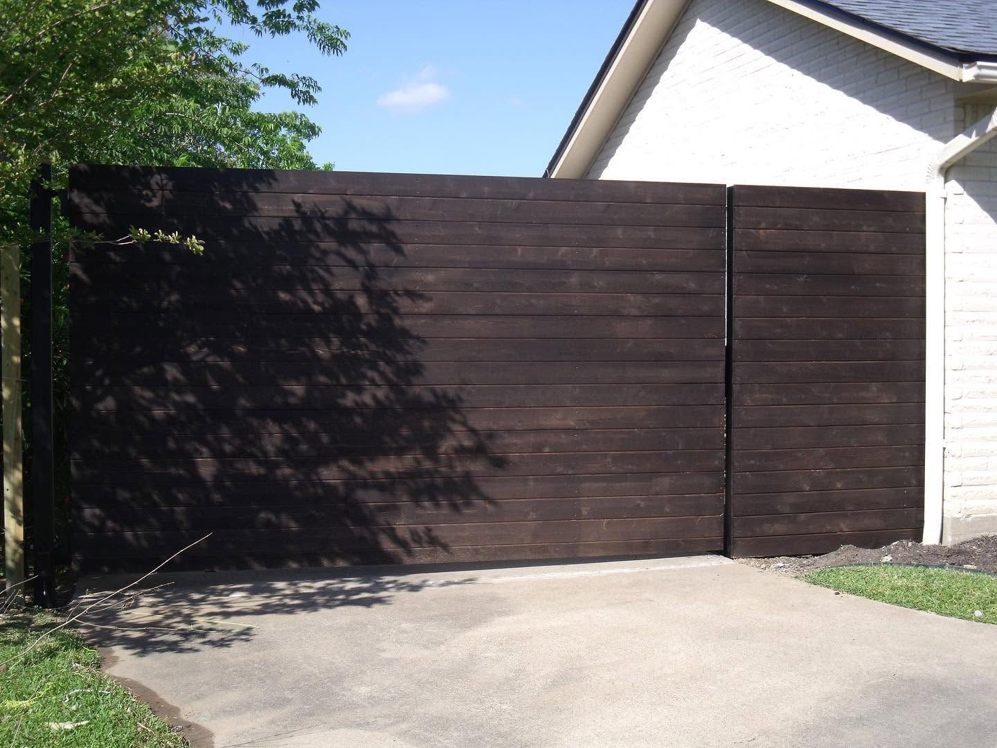Dark wooden driveway gate beside a white house, with sunlit concrete and tree shadows.