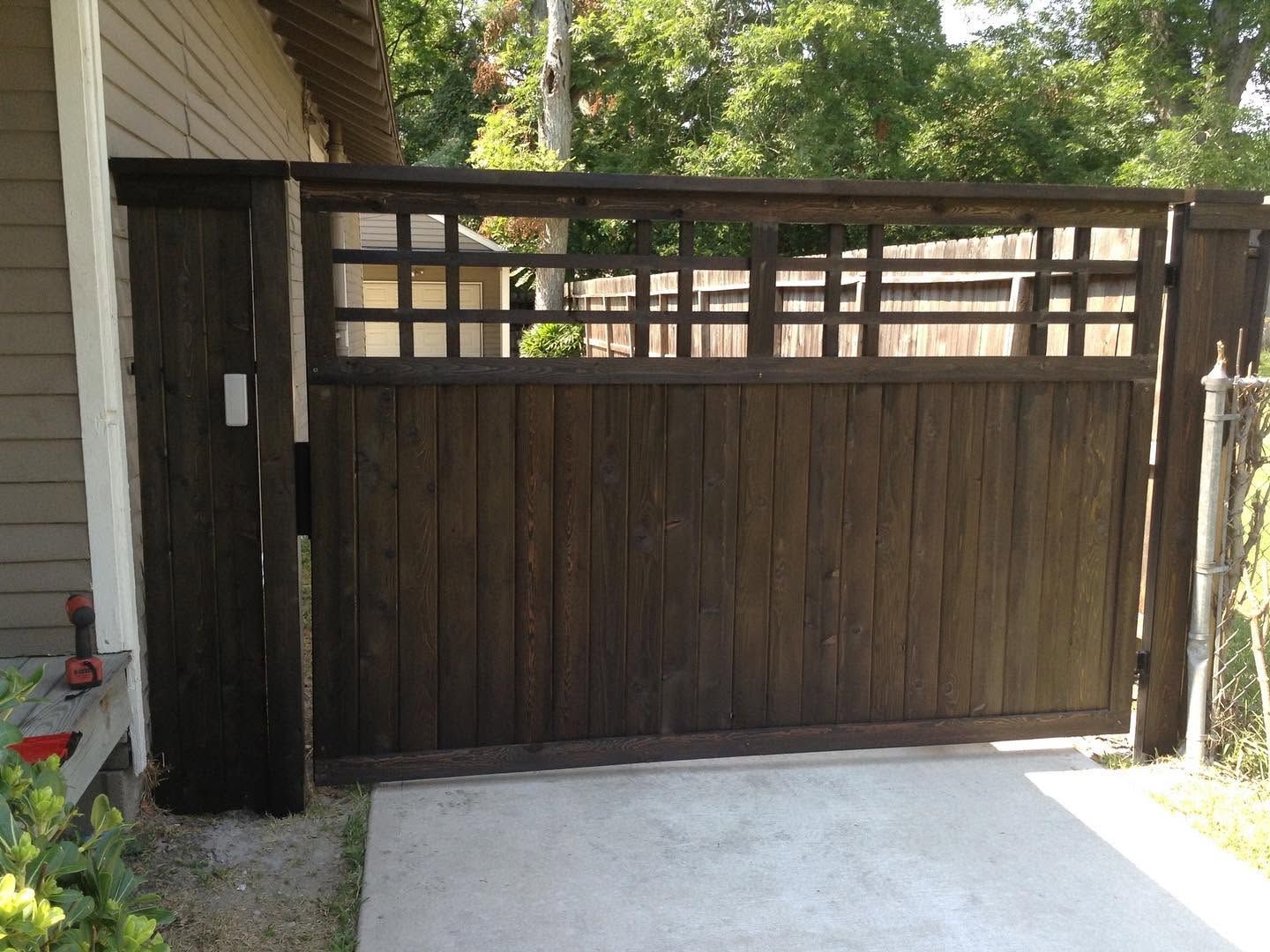 Wooden driveway gate with a dark fence and concrete driveway in front, beside a house and trees