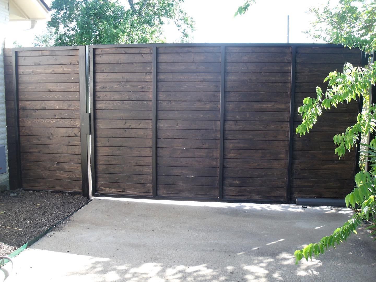 Dark wooden driveway gate beside a concrete path, with greenery in the background