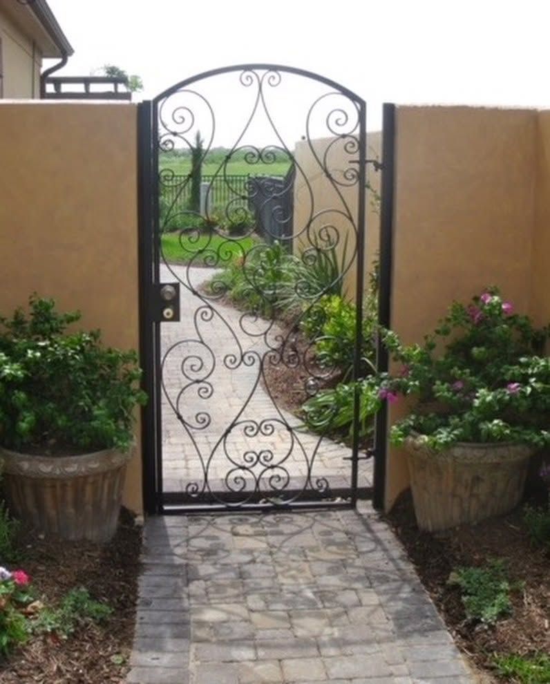 Wrought-iron garden gate between stucco walls, opening to a stone path and lush greenery beyond.