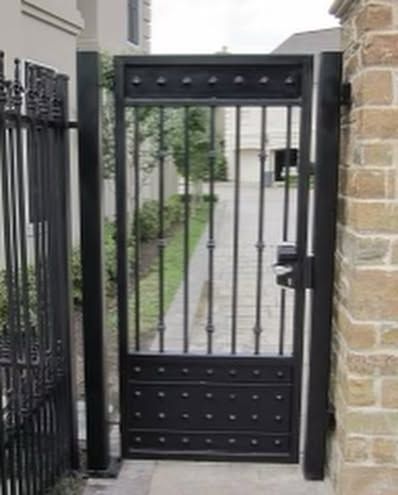 Black metal pedestrian gate beside stone wall, opening onto a paved residential driveway.